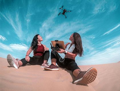 two women on a dubai sand dune control a drone under a bright blue sky.